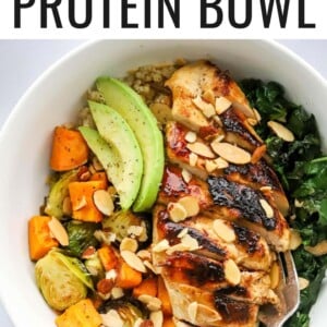 An overhead photo looking down at a chicken protein bowl with roasted veggies, slivered roasted almonds, avocado slices and sautéed kale. A fork rests inside the bowl.