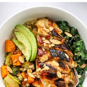 An overhead photo looking down at a chicken protein bowl with roasted veggies, slivered roasted almonds, avocado slices and sautéed kale. A fork rests inside the bowl.