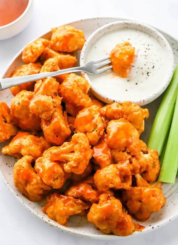 A plate of air fryer buffalo cauliflower with a bowl of ranch and celery sticks. A fork has a piece of cauliflower dipped into the ranch.