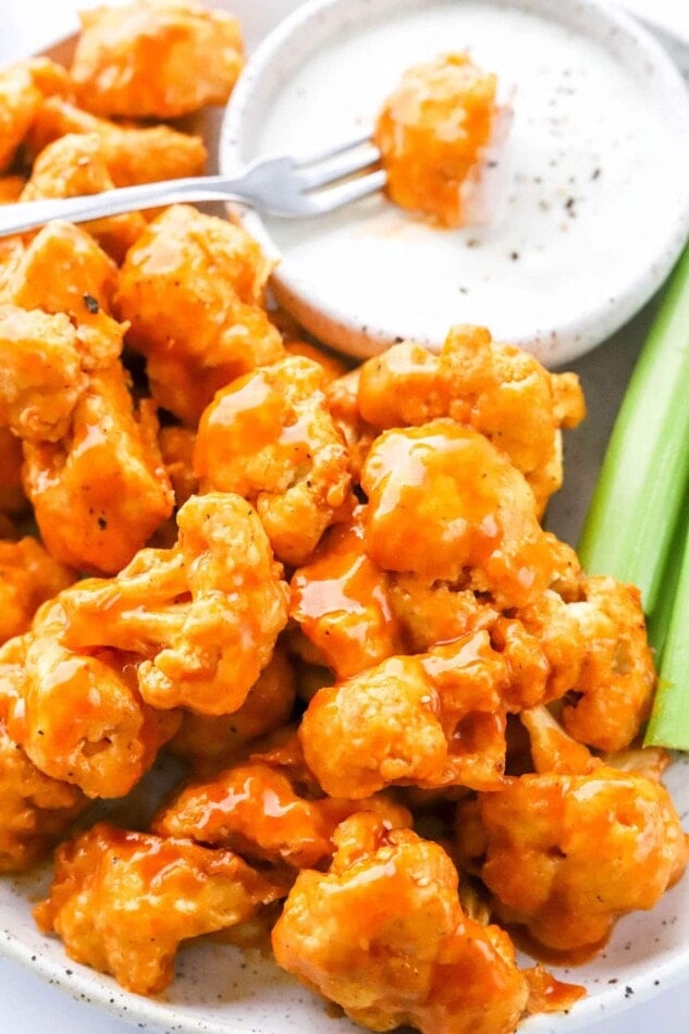 A close up of air fryer buffalo cauliflower on a plate with a bowl of ranch and celery. A fork is dipping a piece of cauliflower into the ranch.