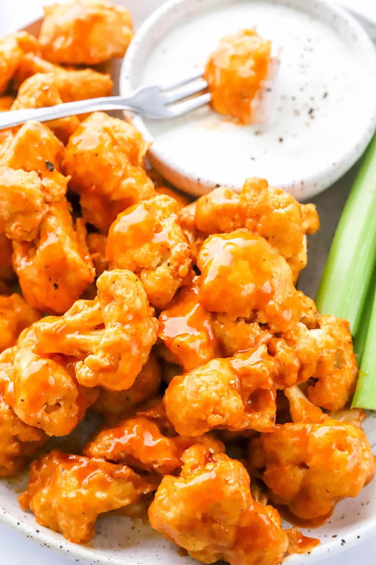 A close up of air fryer buffalo cauliflower on a plate with a bowl of ranch and celery. A fork is dipping a piece of cauliflower into the ranch.
