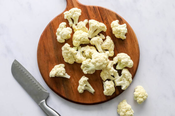 Cauliflower florets on a wooden cutting board. A knife is laying next to the cutting board.