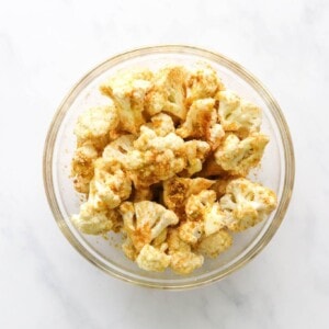 Cauliflower florets tossed in seasoning in a glass mixing bowl.