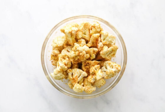 Cauliflower florets tossed in seasoning in a glass mixing bowl.