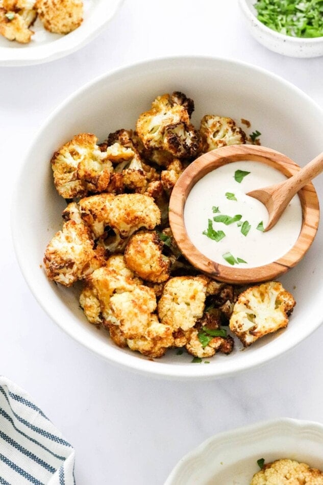 A bowl containing air fried cauliflower. There is a smaller bowl resting inside the larger bowl with some ranch dip and a small wooden spoon.