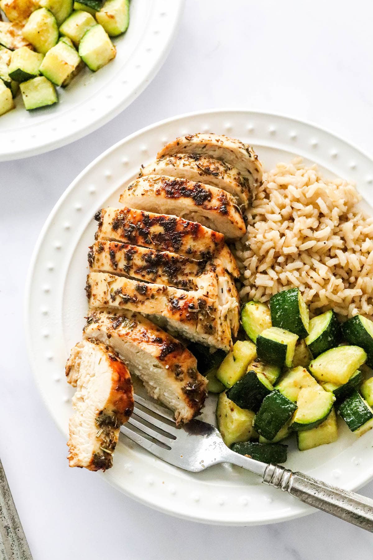 An air fryer chicken breast sliced and served on a plate with brown rice and zucchini. A fork rests on the plate with a piece of chicken.