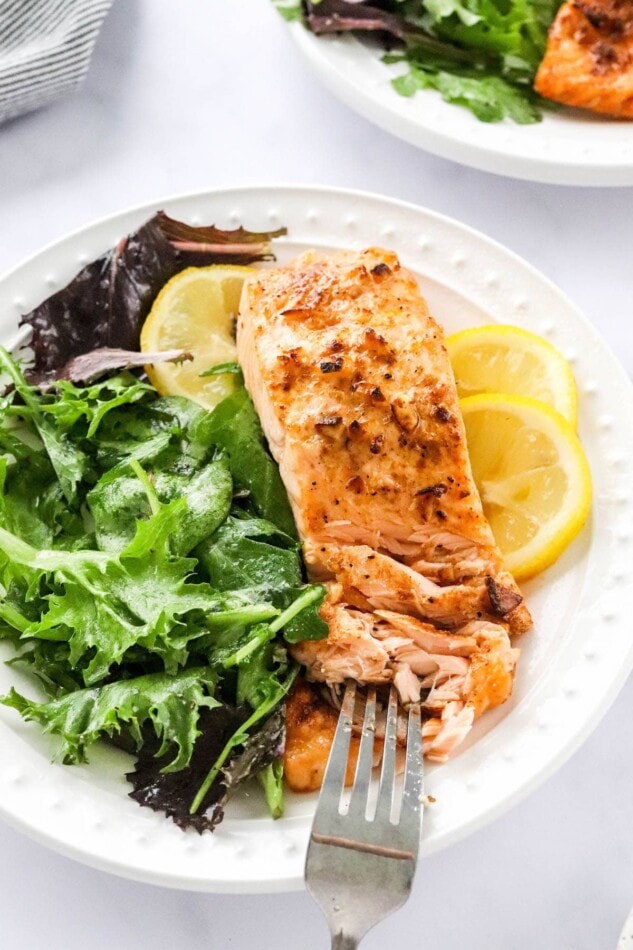 A piece of air fryed salmon on a plate served with leafy greens and lemon slices. A fork is pulling the salmon apart.