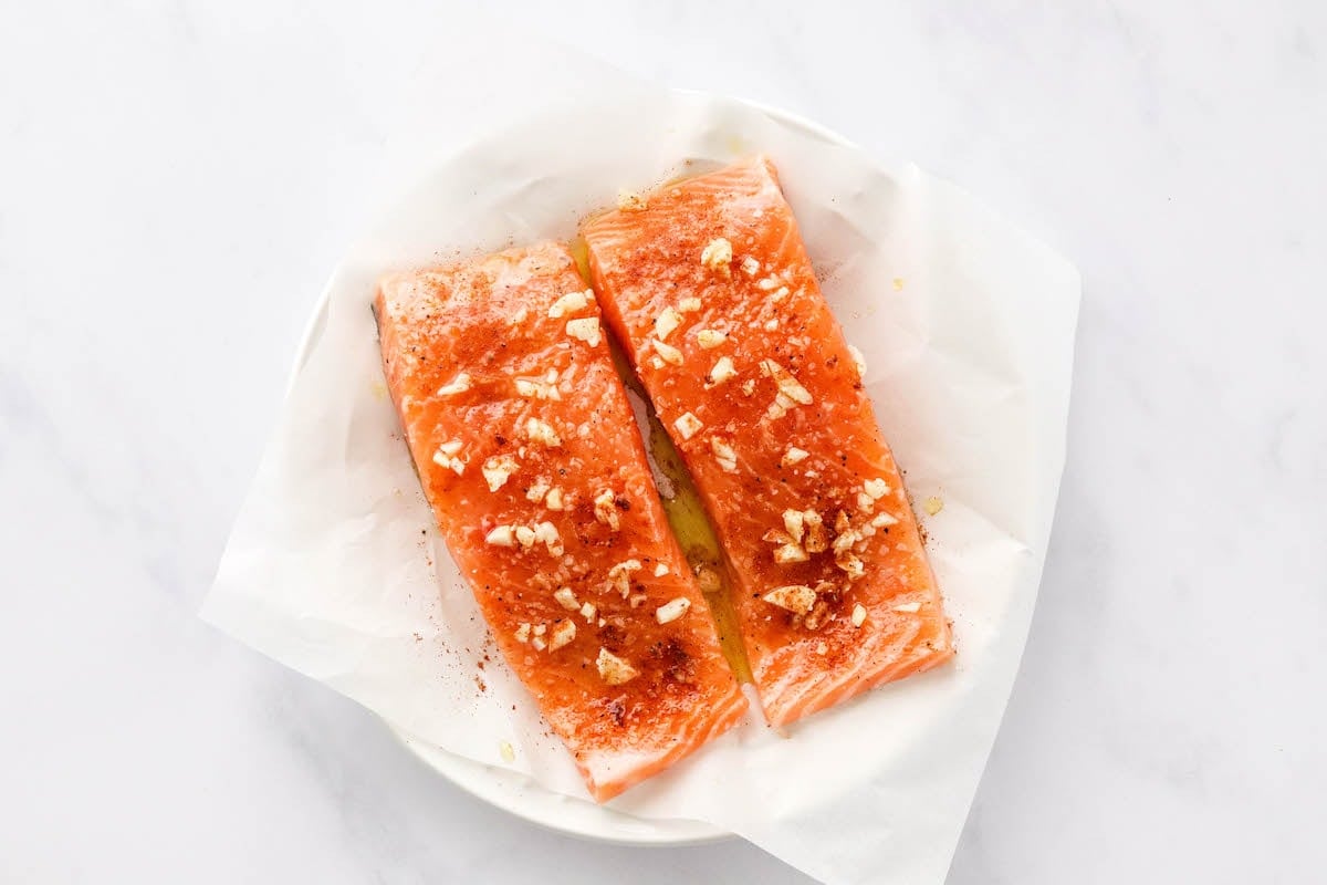 Two seasoned salmon filets on a piece of parchment paper on top of a plate.
