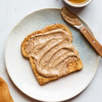 A slice of toast slathered with almond butter on a plate. There is a cup of tea next to the plate.