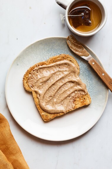 A slice of toast slathered with almond butter on a plate. There is a cup of tea next to the plate.