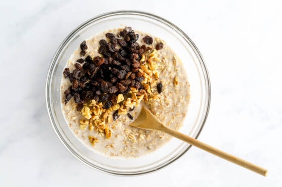 Banana baked oatmeal in a glass mixing bowl. Raisins and walnuts have been added to the bowl but not yet mixed in. A wooden spoon rests in the bowl.