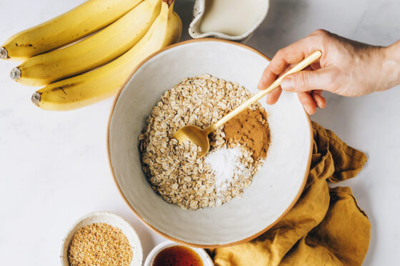 Oats, baking powder, cinnamon, and salt in a mixing bowl. A hand is using a gold spoon to mix the ingredients.