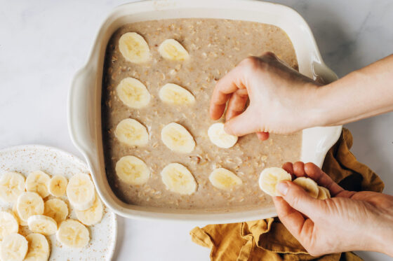 The banana bread baked oatmeal mixture has been poured into the 8x8 baking dish and a hand is placing slices of banana in a 4x4 pattern.