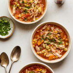 Three bowls of Brunswick stew. A bottle of hot sauce as well as a small bowl of fresh parsley are nearby for topping alongside two metal spoons.