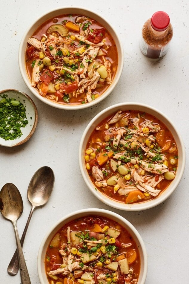 Three bowls of Brunswick stew. A bottle of hot sauce as well as a small bowl of fresh parsley are nearby for topping alongside two metal spoons.