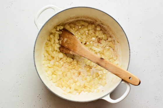 A dutch oven with onions and garlic sautéing in olive oil. A wooden spoon rests in the pot.