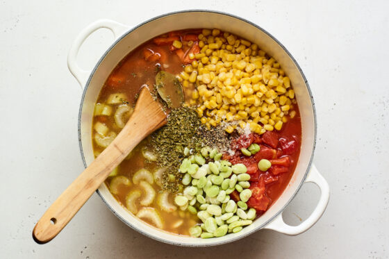 A dutch oven with carrots, celery and potatoes, canned tomatoes, broth, corn, lima beans, worcestershire sauce, parsley, bay leaf, salt, pepper and hot sauce added to it.