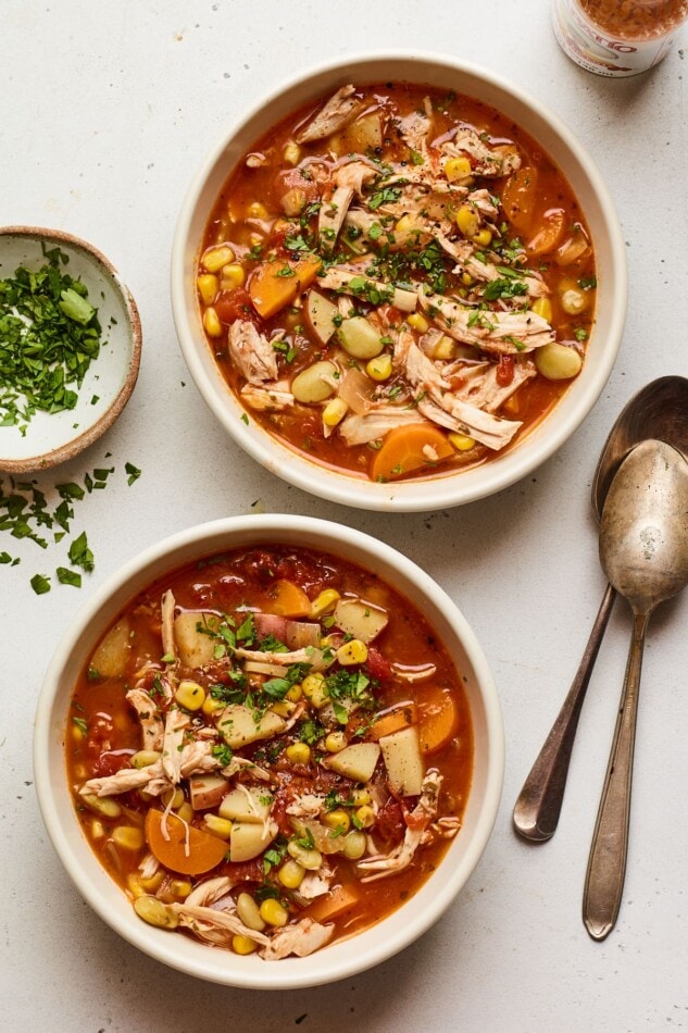 Two bowls containing healthy Brunswick stew. There are two metal spoons next to the bowls as well as a small dish with fresh parsley.