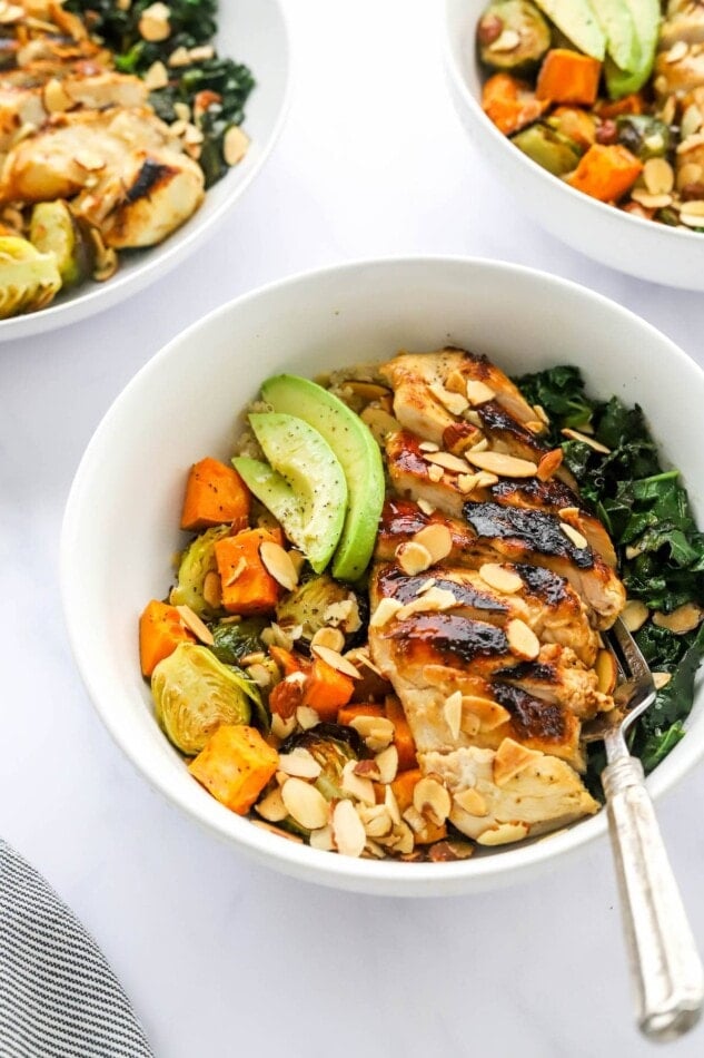A chicken protein bowl with roasted veggies, slivered roasted almonds, avocado slices and sautéed kale. A fork rests inside the bowl.