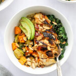 An overhead photo looking down at a chicken protein bowl with roasted veggies, slivered roasted almonds, avocado slices and sautéed kale. A fork rests inside the bowl.