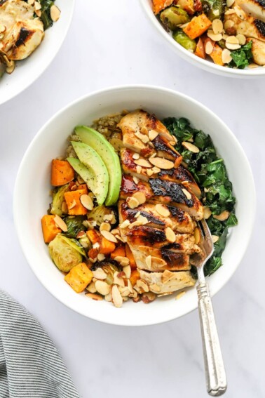 An overhead photo looking down at a chicken protein bowl with roasted veggies, slivered roasted almonds, avocado slices and sautéed kale. A fork rests inside the bowl.