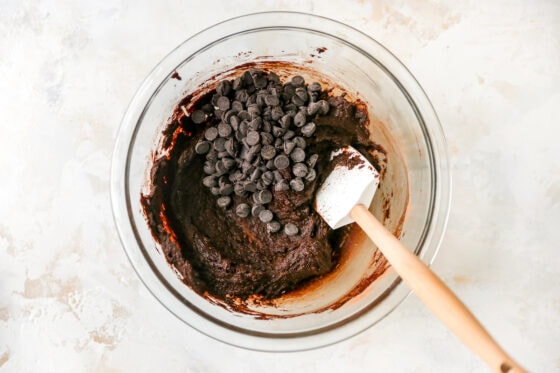 Chocolate chips added to chocolate peppermint cookie dough in a glass mixing bowl. A spatula rests in the bowl.