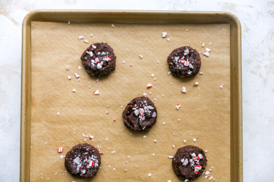 Chocolate peppermint cookies topped with candy cane pieces on a baking sheet lined with brown parchment paper.