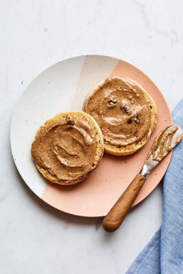 A plate with two halves of an english muffin, slathered with homemade cinnamon raisin almond butter.