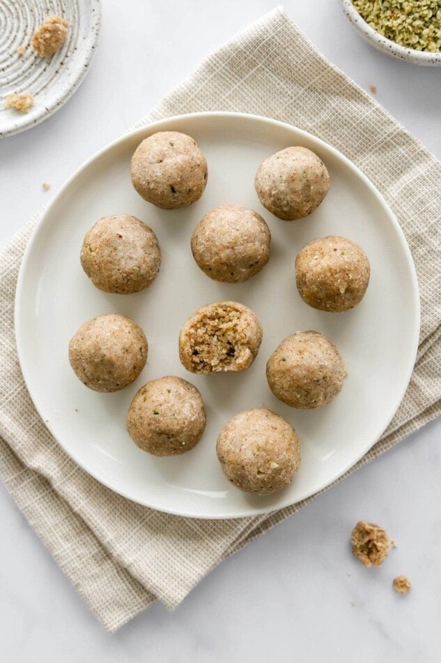 An overhead photo looking down on 10 coconut protein balls on a plate. The center ball has a bite taken out of it. The plate is on a khaki dish cloth.