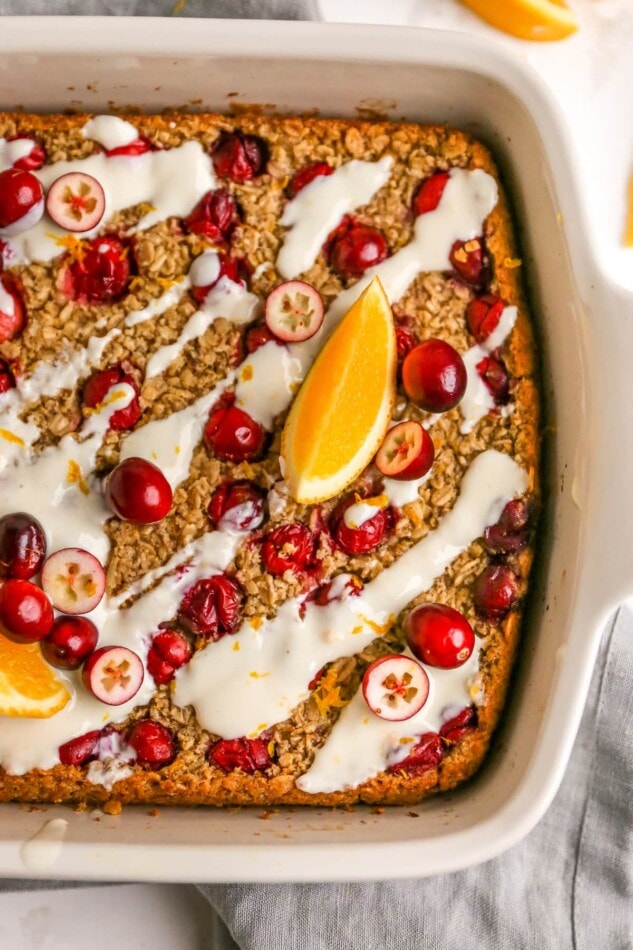 An overhead photo looking down at a baking dish containing cranberry orange baked oatmeal. There are whole and halved cranberries sprinkled on top next to orange slices and the oatmeal has been coated in a glaze.