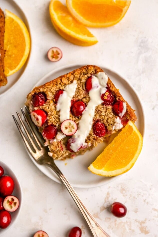 An overhead photo of a plate containing a slice of cranberry orange baked oatmeal. A fork is resting next to the slice on the plate Orange slices and cranberries are scattered around.