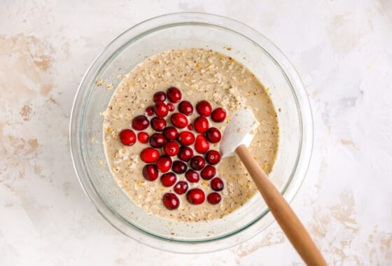 A mixing bowl containing cranberry orange oatmeal mixture. Whole cranberries have been added to the bowl. A spatula is ready to mix the ingredients together.