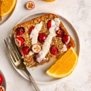 An overhead photo of a plate containing a slice of cranberry orange baked oatmeal. A fork is resting next to the slice on the plate Orange slices and cranberries are scattered around.