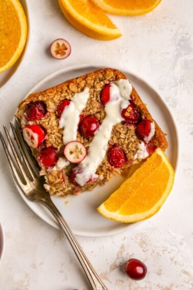 An overhead photo of a plate containing a slice of cranberry orange baked oatmeal. A fork is resting next to the slice on the plate Orange slices and cranberries are scattered around.