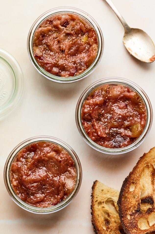 An overhead photo looking down at 3 jars of homemade fig jam. A spoon and two slices of toasted bread are next to the jars.