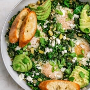 A skillet with green shakshuka that has been topped with slices of avocado and toasted bread.