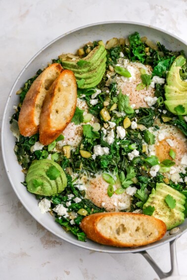 A skillet with green shakshuka that has been topped with slices of avocado and toasted bread.