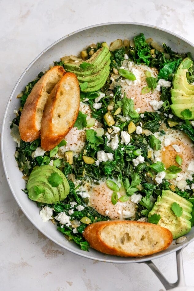 A skillet with green shakshuka that has been topped with slices of avocado and toasted bread.