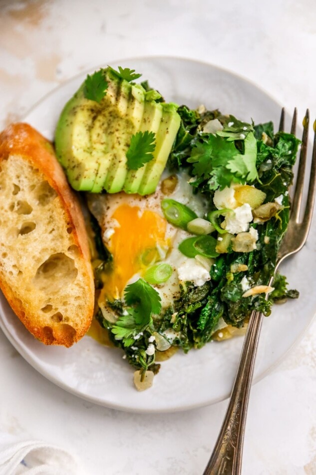 A closeup photo of a plate containing a serving of green shakshuka with a slice of toasted bread and some sliced avocado. A fork rests on the plate.