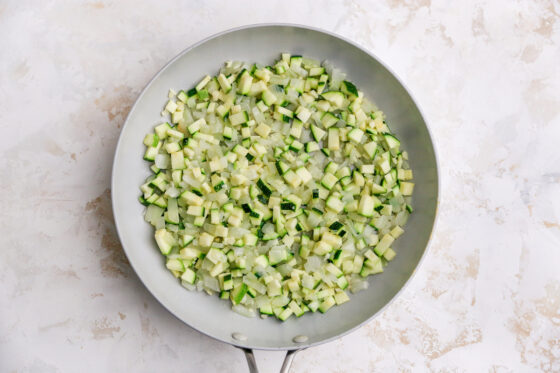 A skillet with onions, zucchini and garlic cooking in oil.