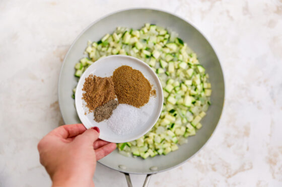 Cumin, coriander, salt and pepper being added to the skillet containing zucchini, onion and garlic.