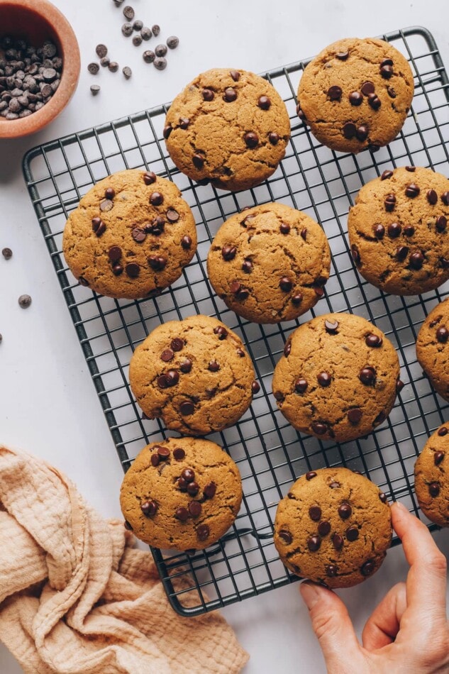 An overhead photo looking down at a wire cooling rack with healthy chocolate chip cookies scattered around it. A hand is removing one of the cookies from the bottom right hand corner.