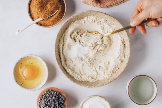 A mixing bowl with flour, baking soda and sea salt. A hand is using a golden spoon to mix the ingredients together.