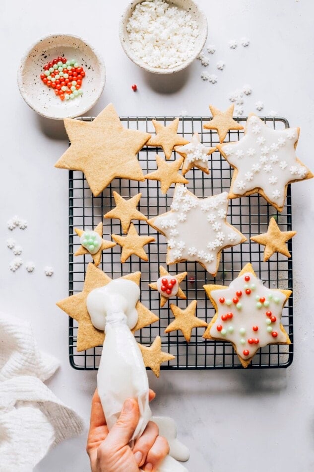 A wire rack with various sized star shaped cookies that are being decorated. There are small dishes around the wire rack with assorted sprinkles. A hand is apply icing to one of the cookies.