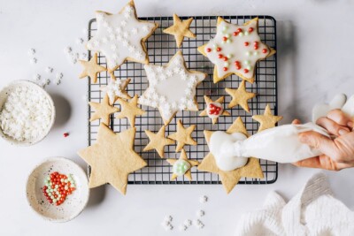 A wire rack with various sized star shaped cookies that are being decorated. There are small dishes around the wire rack with assorted sprinkles. A hand is apply icing to one of the cookies.