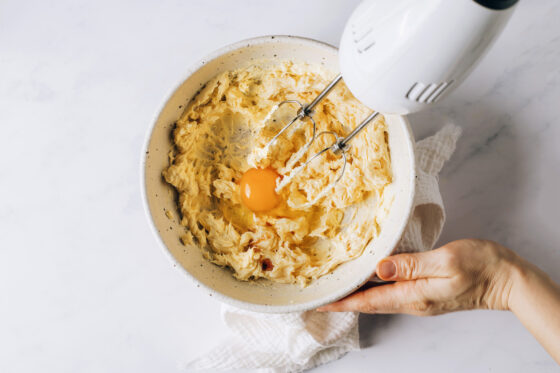 An egg and vanilla extract added to a bowl of creamed butter and maple syrup. A hand mixer is ready to mix them together.