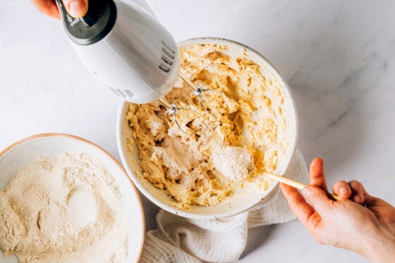 Flour mixture being added to the egg, vanilla, butter and maple syrup mixture by a spoon. The hand mixer is in the bowl.