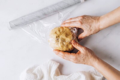 A hand is using saran wrap to wrap up a ball of sugar cookie dough.