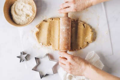 Two hands using a small rolling pin to roll out healthy sugar cookie dough onto parchment paper. Two star shaped cookie cutters are nearby as well as a bowl of extra flour.