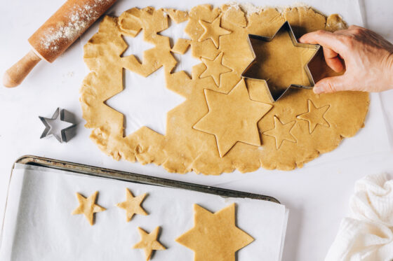 A hand using a star shaped cookie cutter to cut out cookies from rolled dough. The cookies are being placed onto a parchment lined baking sheet.